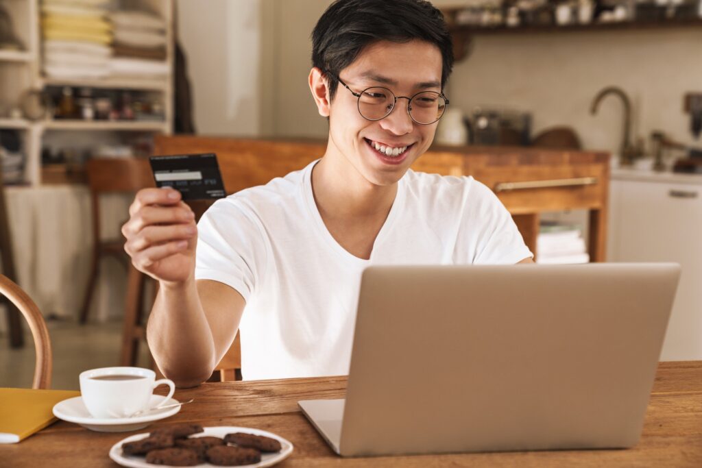 Image of smiling asian man holding credit card while using laptop