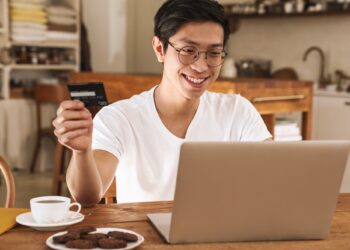 Image of smiling asian man holding credit card while using laptop