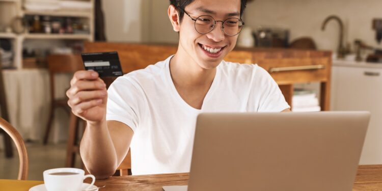 Image of smiling asian man holding credit card while using laptop