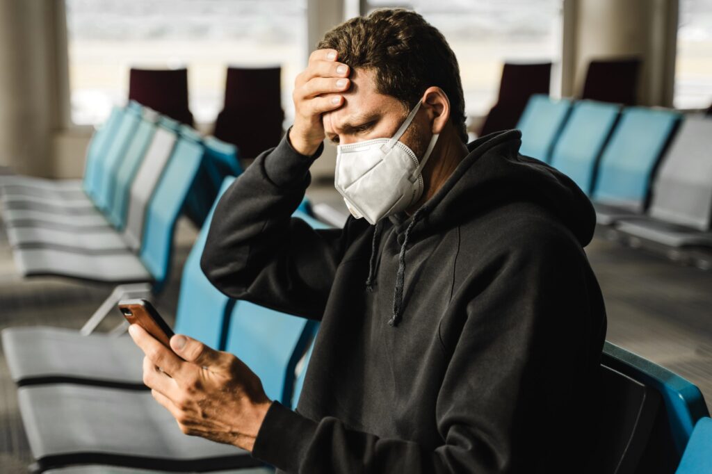 Men in respirator mask sitting at the airport, measuring temperature with palm and using mobile phon