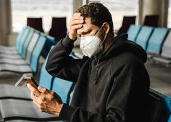 Men in respirator mask sitting at the airport, measuring temperature with palm and using mobile phon