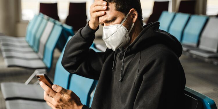 Men in respirator mask sitting at the airport, measuring temperature with palm and using mobile phon