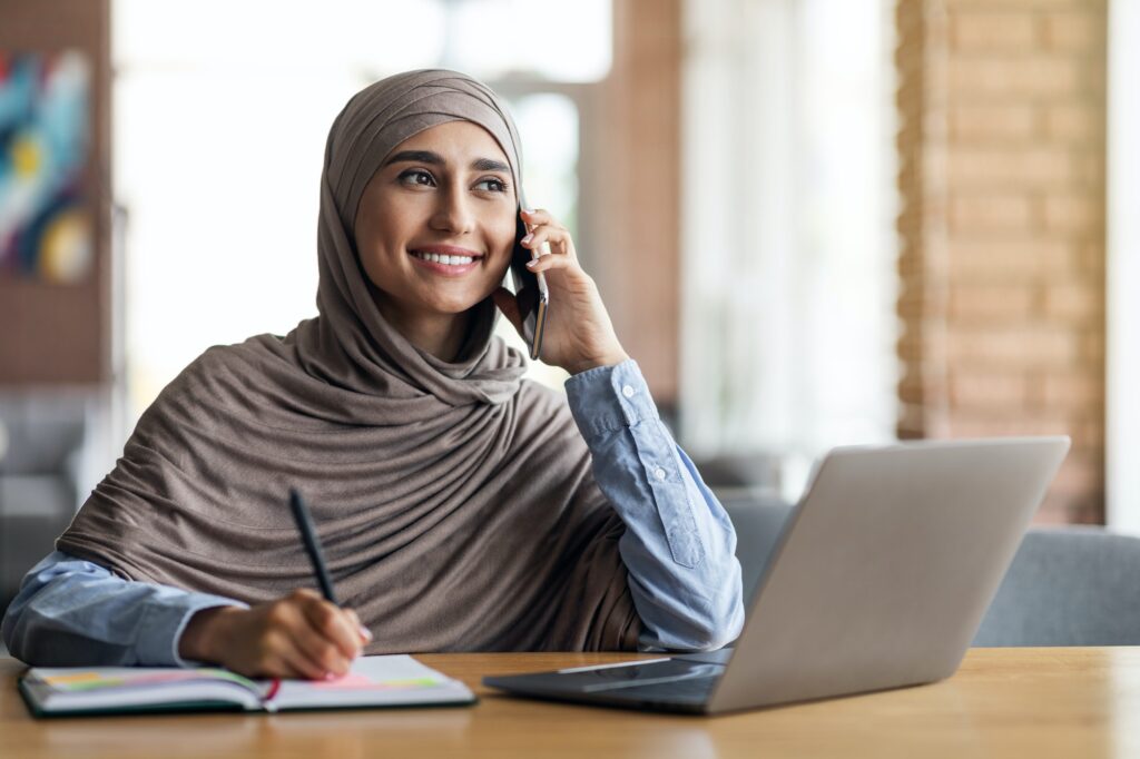Muslim woman freelancer talking to client on phone, taking notes
