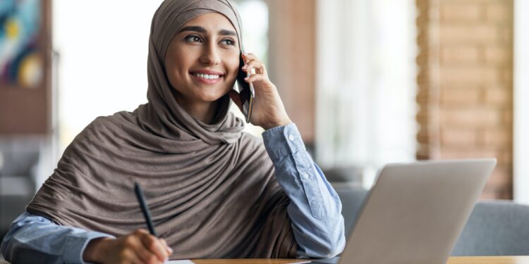 Muslim woman freelancer talking to client on phone, taking notes
