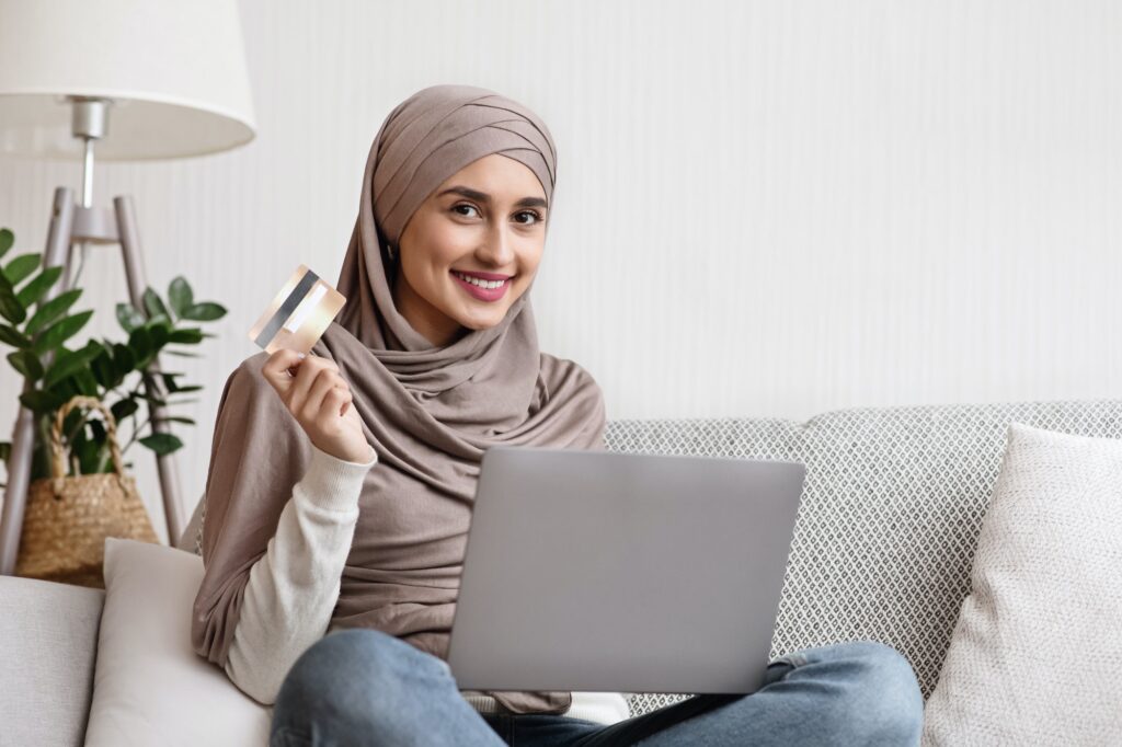 Smiling islamic woman sitting on sofa with laptop and credit card