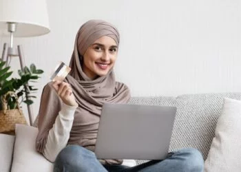 Smiling islamic woman sitting on sofa with laptop and credit card