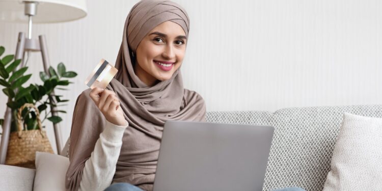 Smiling islamic woman sitting on sofa with laptop and credit card