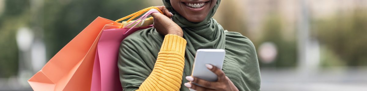 Online Banking. Cheerful Black Muslim Female With Smartphone And Shopping Bags Outdoors